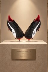 A pair of black and white soccer cleats with red accents and white script logos, displayed on a beige pedestal in a museum or gallery setting.